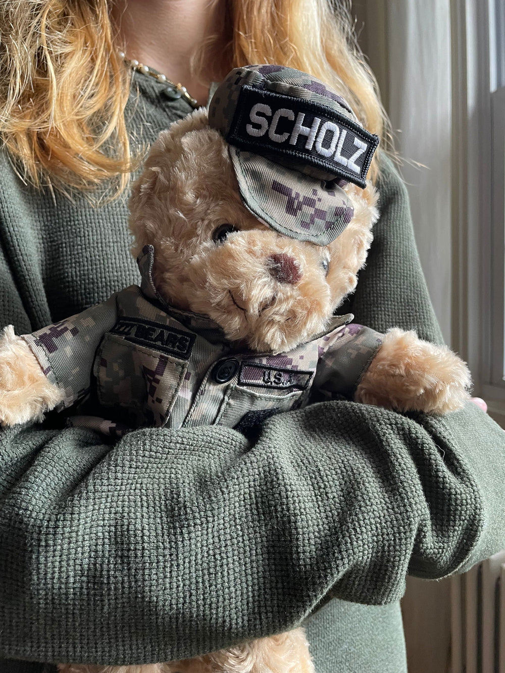 A person in a green sweater holds the Sailor Sleeptight – Navy Teddy Bear in cammo by ZZZ BEARS, complete with storybook and sleep system—ideal for any child’s nighttime routine.